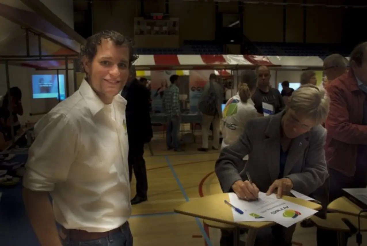 At the same event, a woman leans over a small table to sign an AnnieS document while Robert McConnell, in an AnnieS shirt, stands in the foreground looking toward the camera.