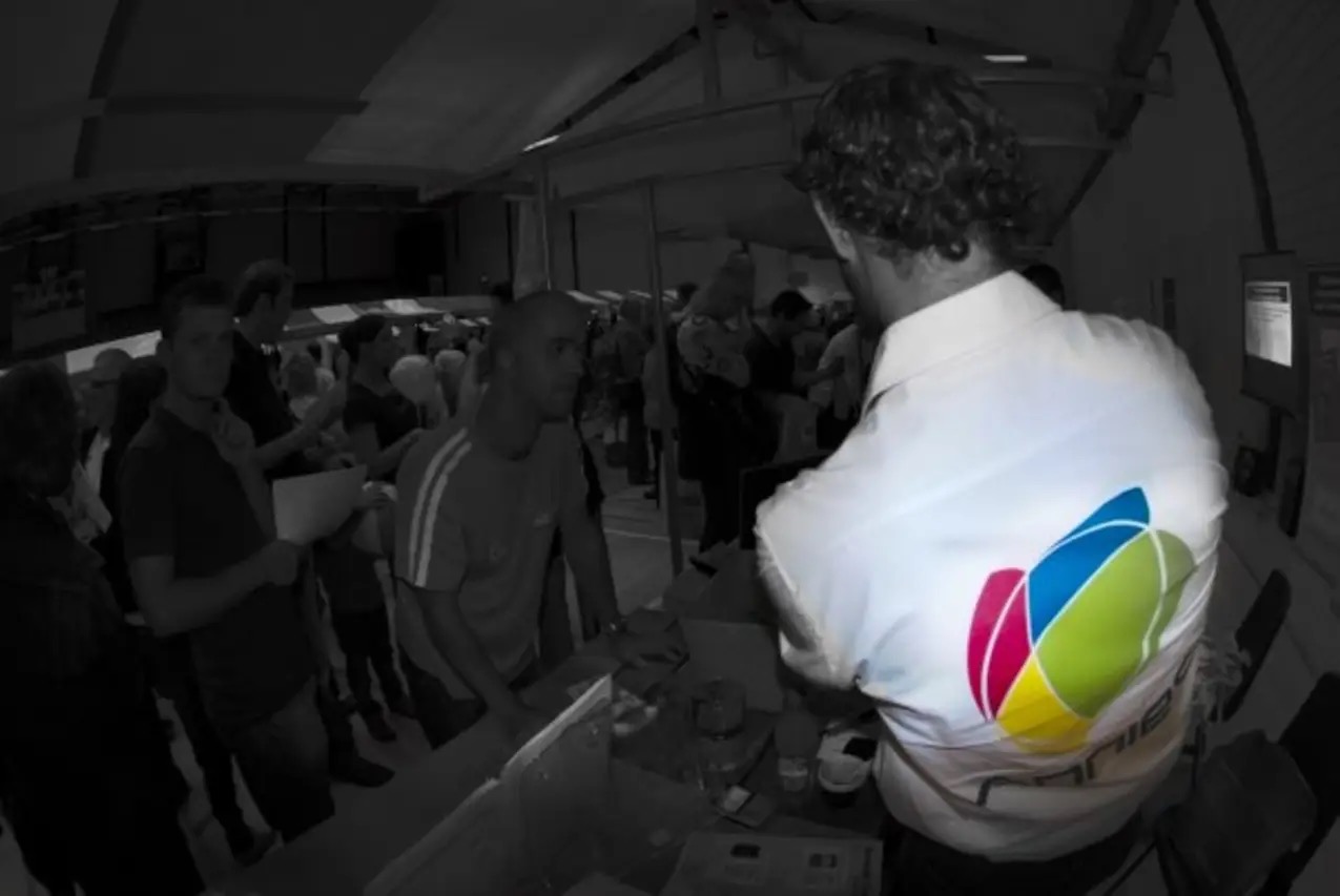 The same AnnieS stand photographed from behind the table and desaturated, so that the multicoloured AnnieS logo on the back of Robert McConnell's shirt is the only colour in an otherwise black-and-white scene of waiting visitors.