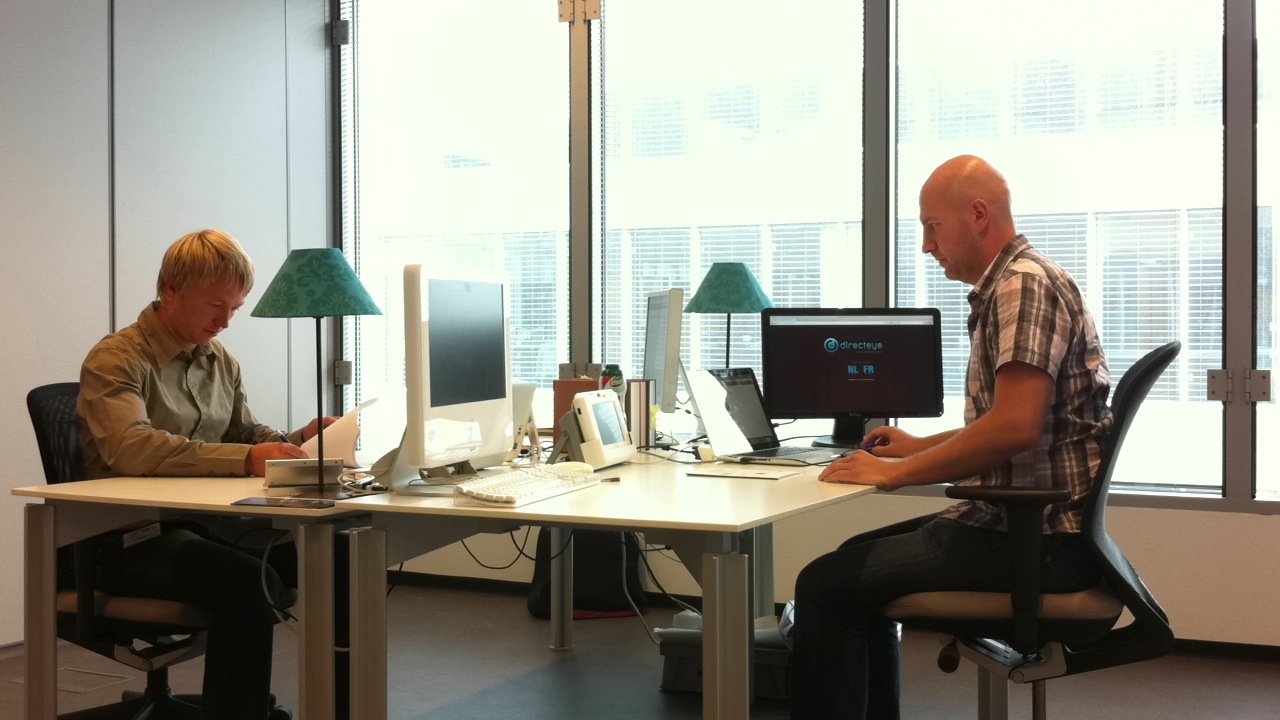 Inside DirectEye’s Ghent office in 2011: two people at facing desks with monitors, a laptop and a desktop video terminal.